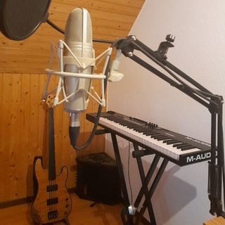 a small bit of my home studio, right under a roof, made of rustic wooden walls. You can see my master keyboard behind a microphone on its perch, and my favorite bass on the back.